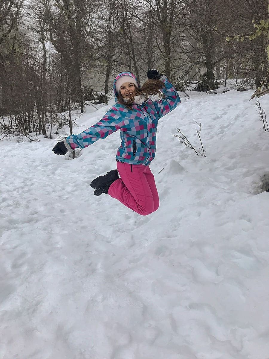 Una chica con campera y pantalón de nieve salta con los brazos en alto en una ladera nevada.