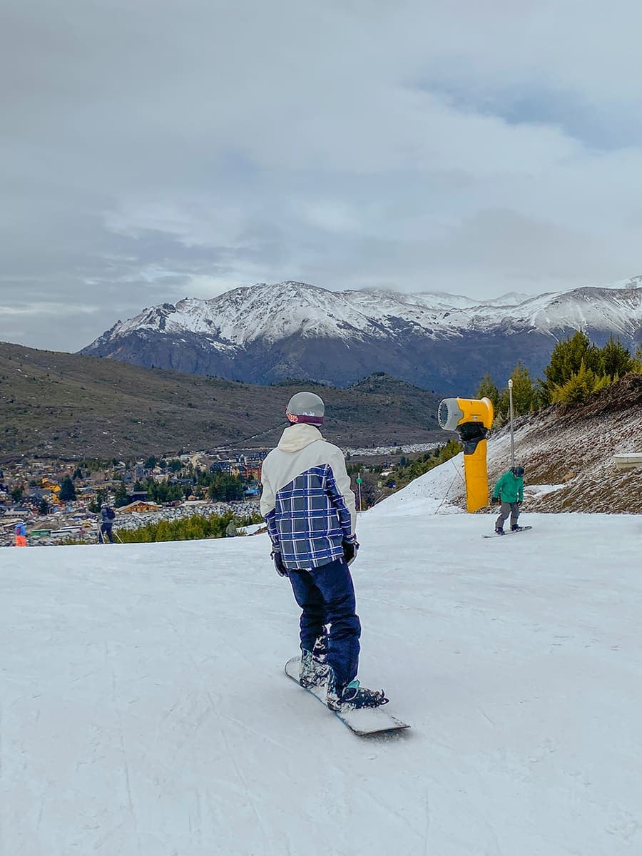 Una persona de espaldas, con tabla de snowboard, mirando Bariloche y el lago desde la pista.